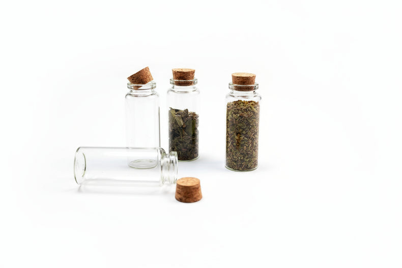 Three small glass bottles with cork lids containing herbs on a dark surface, with a smoldering sage bundle in the background.