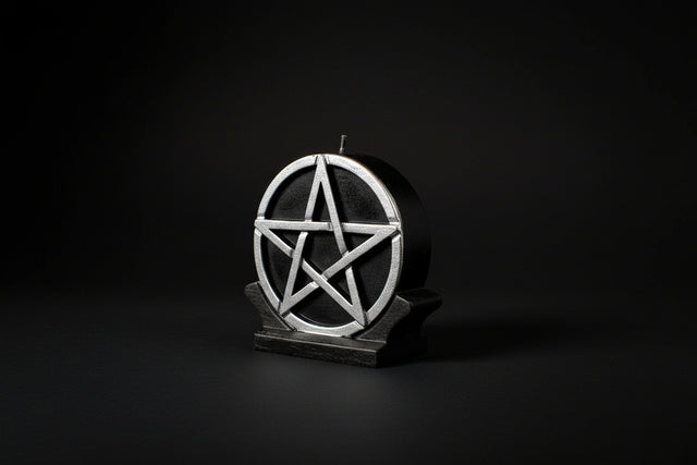A 5-inch black ritual candle with a silver pentacle design on the top, placed on a white surface with a wooden container in the background.