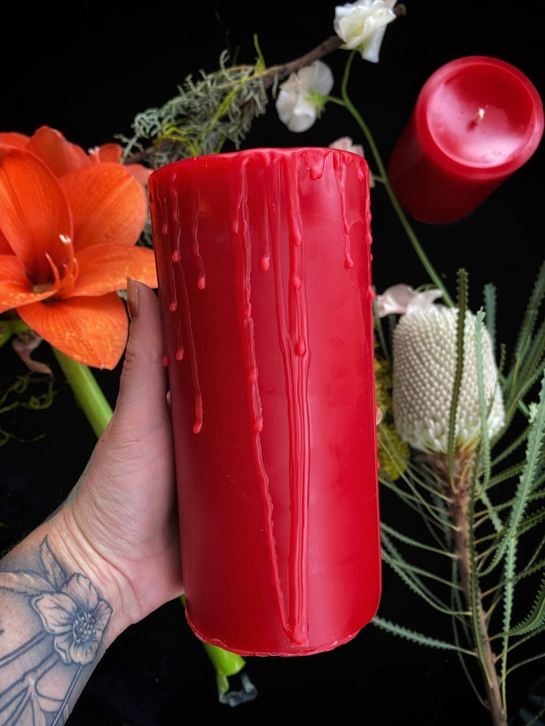 Red candle with a textured surface held by a hand, surrounded by flowers on a dark background