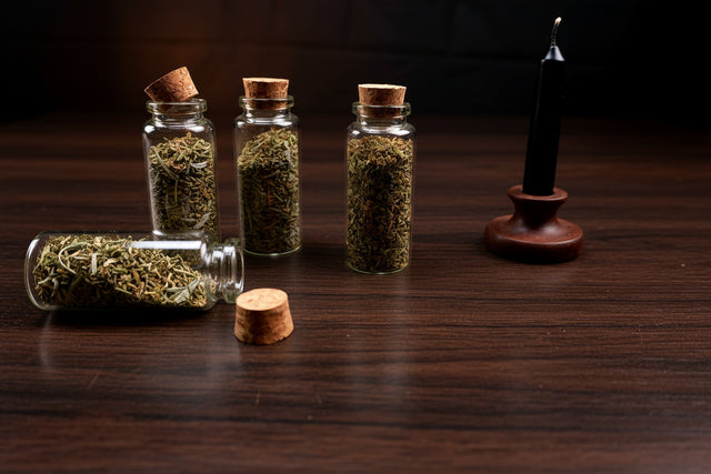 Three small glass bottles with cork lids containing herbs on a dark surface, with a smoldering sage bundle in the background.