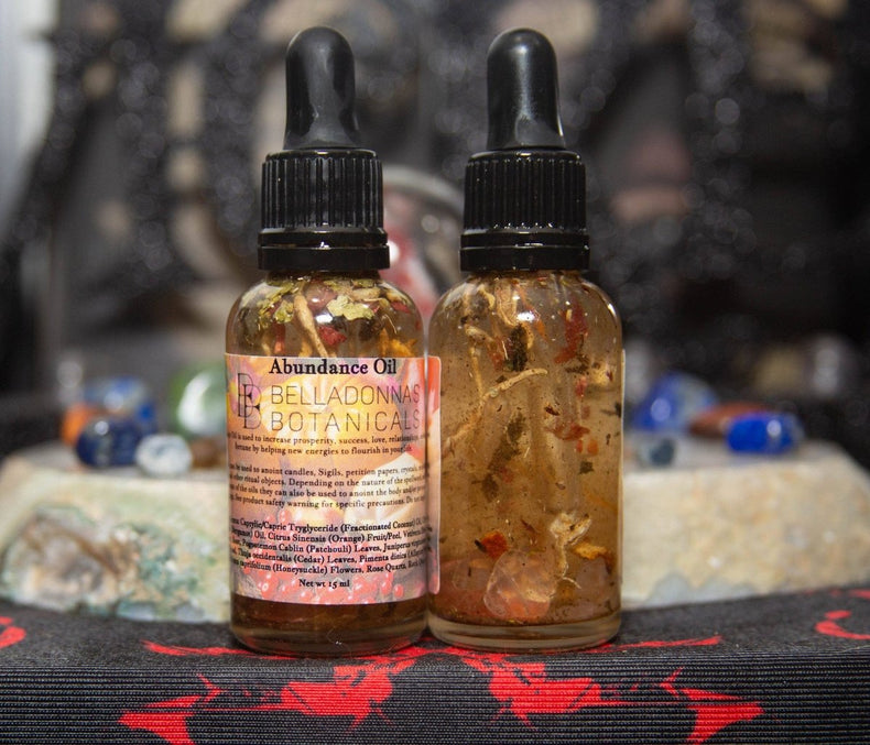 Two bottles of Abundance Oil with botanicals on a dark surface with blurred background