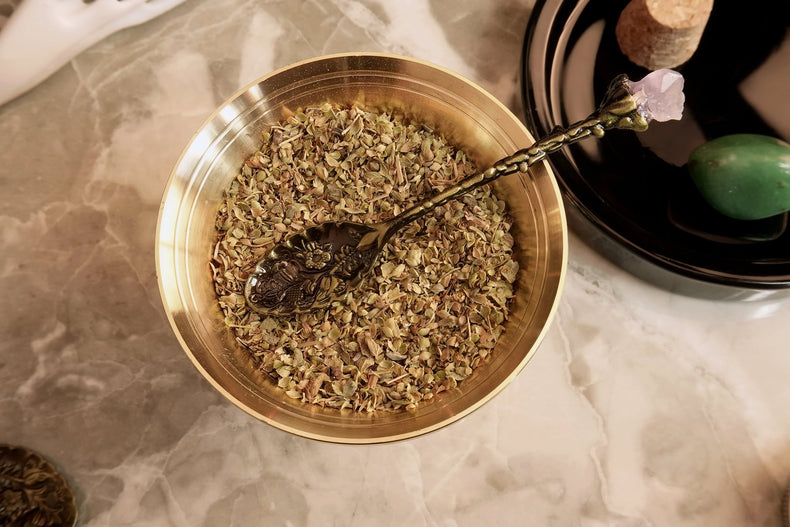 A gold-colored metal spoon with an amethyst cluster handle, resting in a bowl filled with loose amethyst pieces, on a marble surface with a candle and other items in the background.