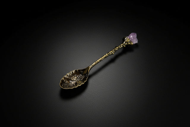 A gold-colored metal spoon with an amethyst cluster handle, resting in a bowl filled with loose amethyst pieces, on a marble surface with a candle and other items in the background.