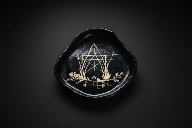 Black embroidered dish with geometric design held by a hand, surrounded by greenery and flowers.