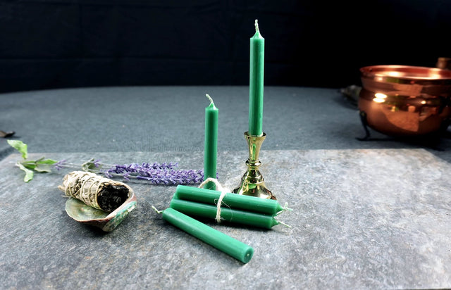 Green chime candles tied with gold string, lit at one end, on a dark background with a copper bowl and herbs.