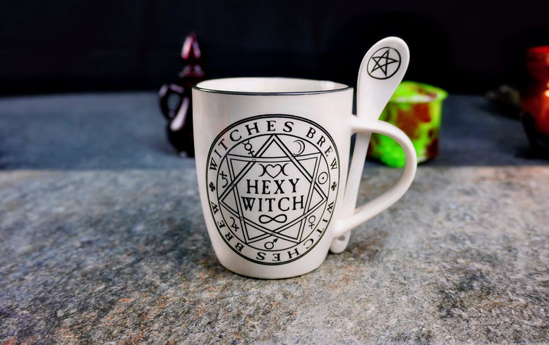 A white ceramic mug with the text 'Hexy Witch' and 'Witches Brew' designs surrounded by occult symbols, accompanied by a matching spoon with a pentacle on the handle, displayed on a kitchen countertop.