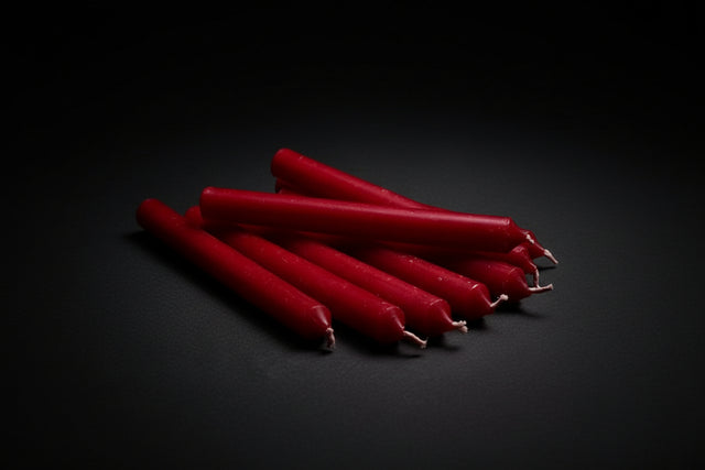 A group of red chime candles arranged on a gray surface with a lighted candle on a gold holder in the background, and a small white and green object to the left.