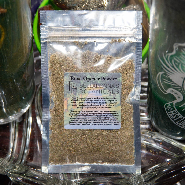 Packaged herb labeled 'Road Opener Powder' by Belladonna's Botanicals on a reflective surface with a candle in the background.