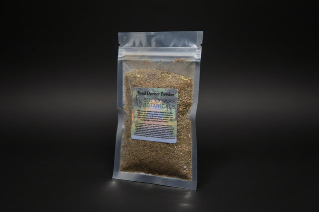 Packaged herb labeled 'Road Opener Powder' by Belladonna's Botanicals on a reflective surface with a candle in the background.