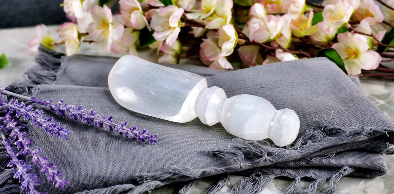 A 6-inch Selenite Athame placed on a gray cloth with purple flowers and a branch of pink flowers in the background.