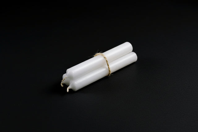 A white ritual chime candle placed on a stone surface, tied with a string, surrounded by purple flowers and a gold-colored bell in the background.