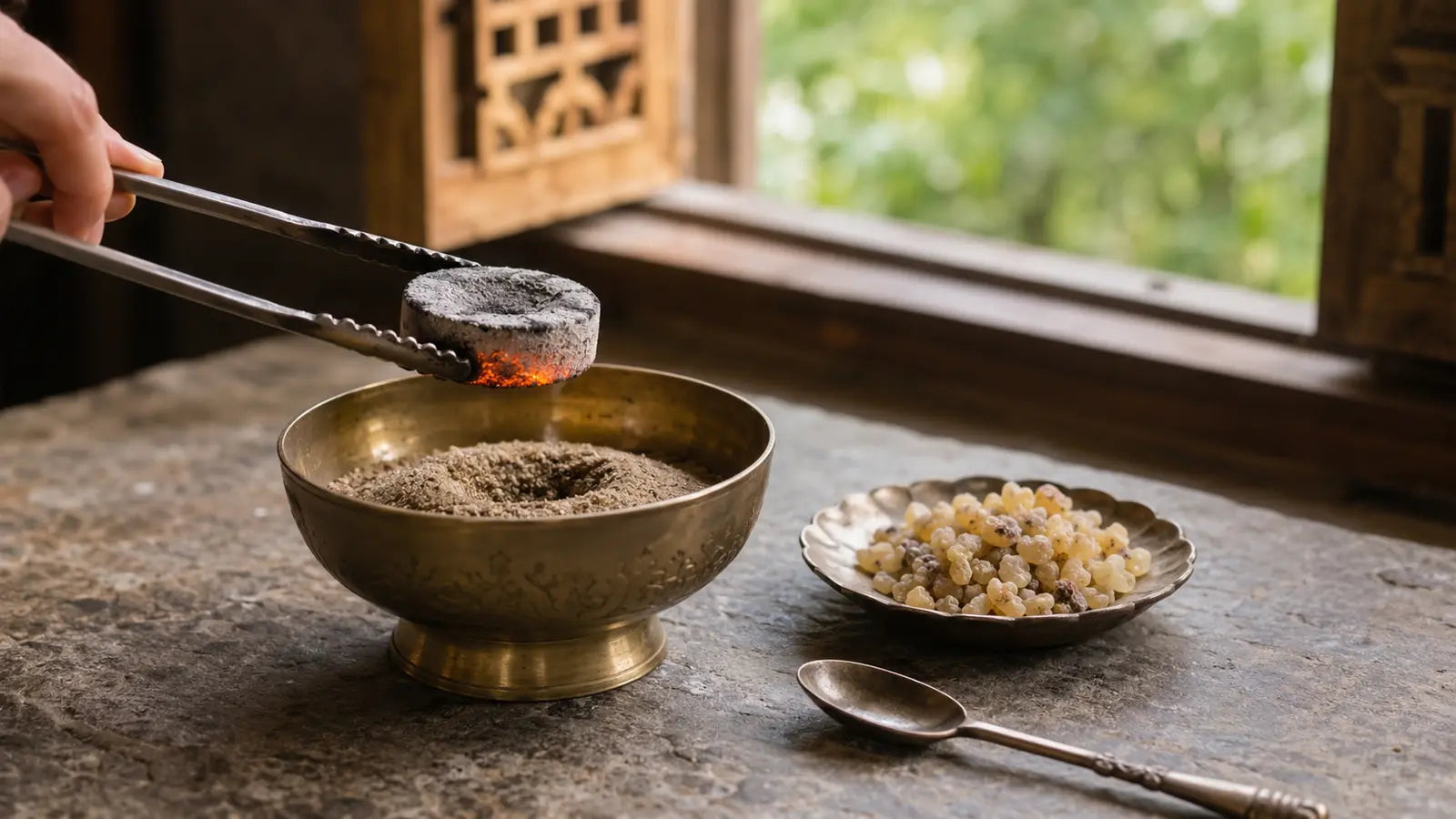 Glowing charcoal disc held with metal tongs above a sand-filled brass bowl beside frankincense resin