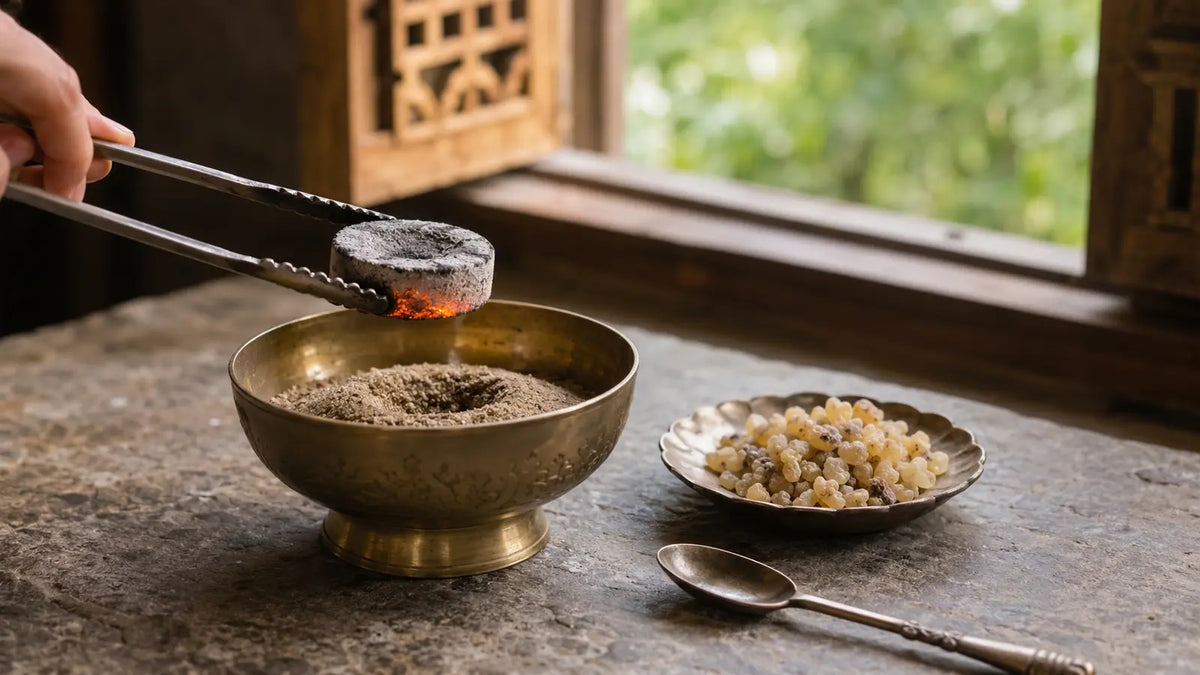 Glowing charcoal disc held with metal tongs above a sand-filled brass bowl beside frankincense resin