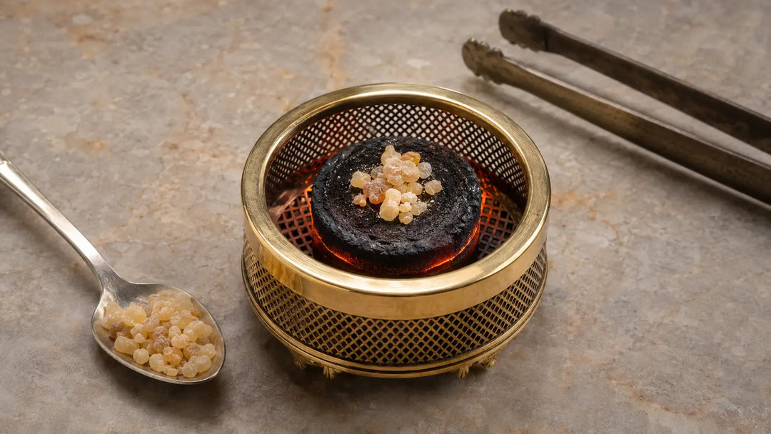 Small measured pinch of frankincense resin on a glowing charcoal disc inside a brass mesh burner with spoon and tongs