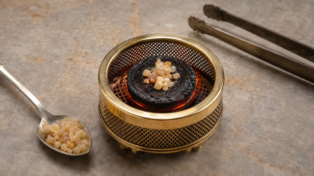 Small measured pinch of frankincense resin on a glowing charcoal disc inside a brass mesh burner with spoon and tongs