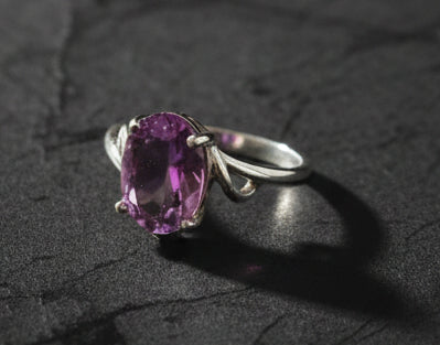 A silver ring with a small oval purple gemstone displayed on a black ring stand against a gray background.