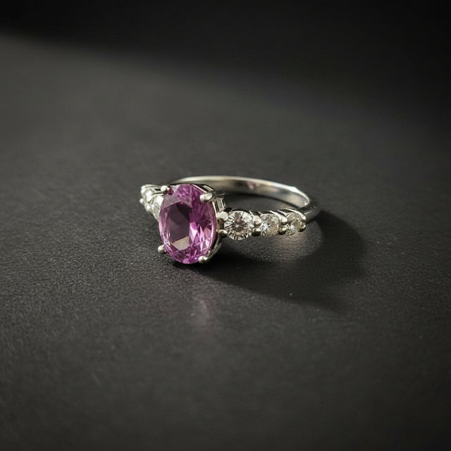 A silver ring with a prominent oval purple gemstone and small clear accent stones, displayed on a black ring stand against a gray background.