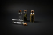 Three small glass bottles with cork lids containing herbs on a dark surface, with a smoldering sage bundle in the background.