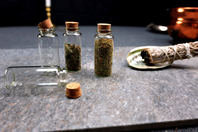 Three small glass bottles with cork lids containing herbs on a dark surface, with a smoldering sage bundle in the background.