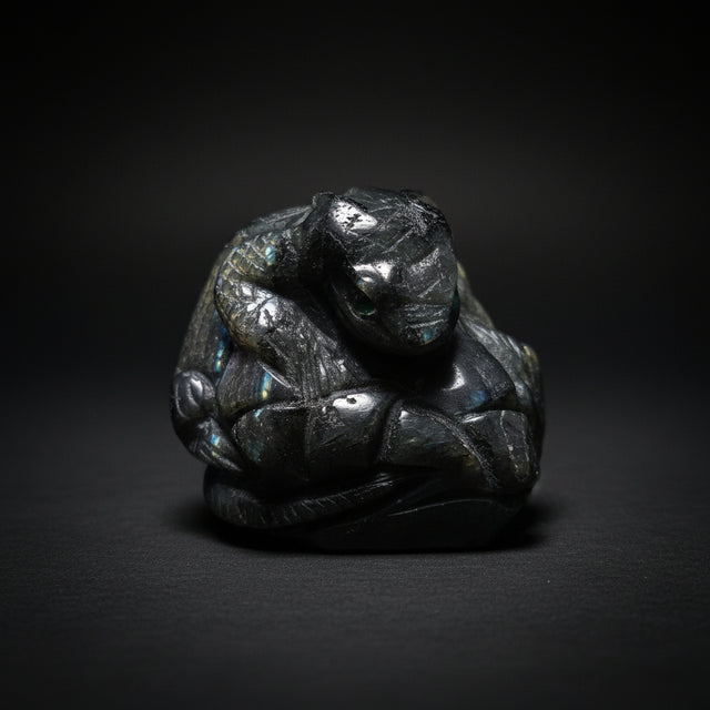 A carved black labradorite figurine of a lizard with a green eye, photographed on a light stone surface.
