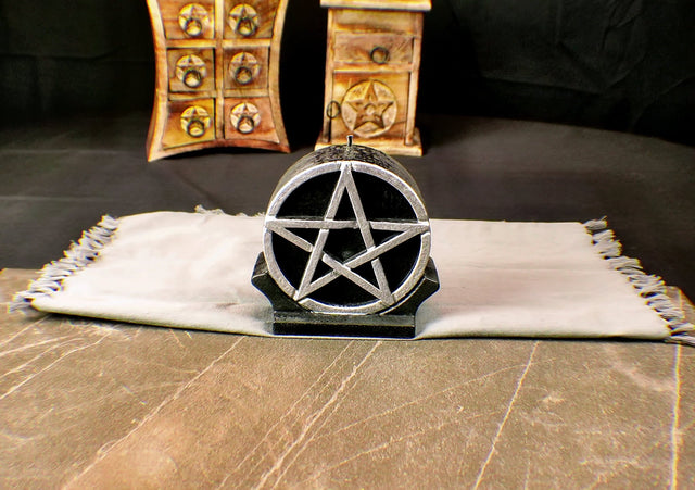 A 5-inch black ritual candle with a silver pentagram design on the top, placed on a white surface with a wooden container in the background.