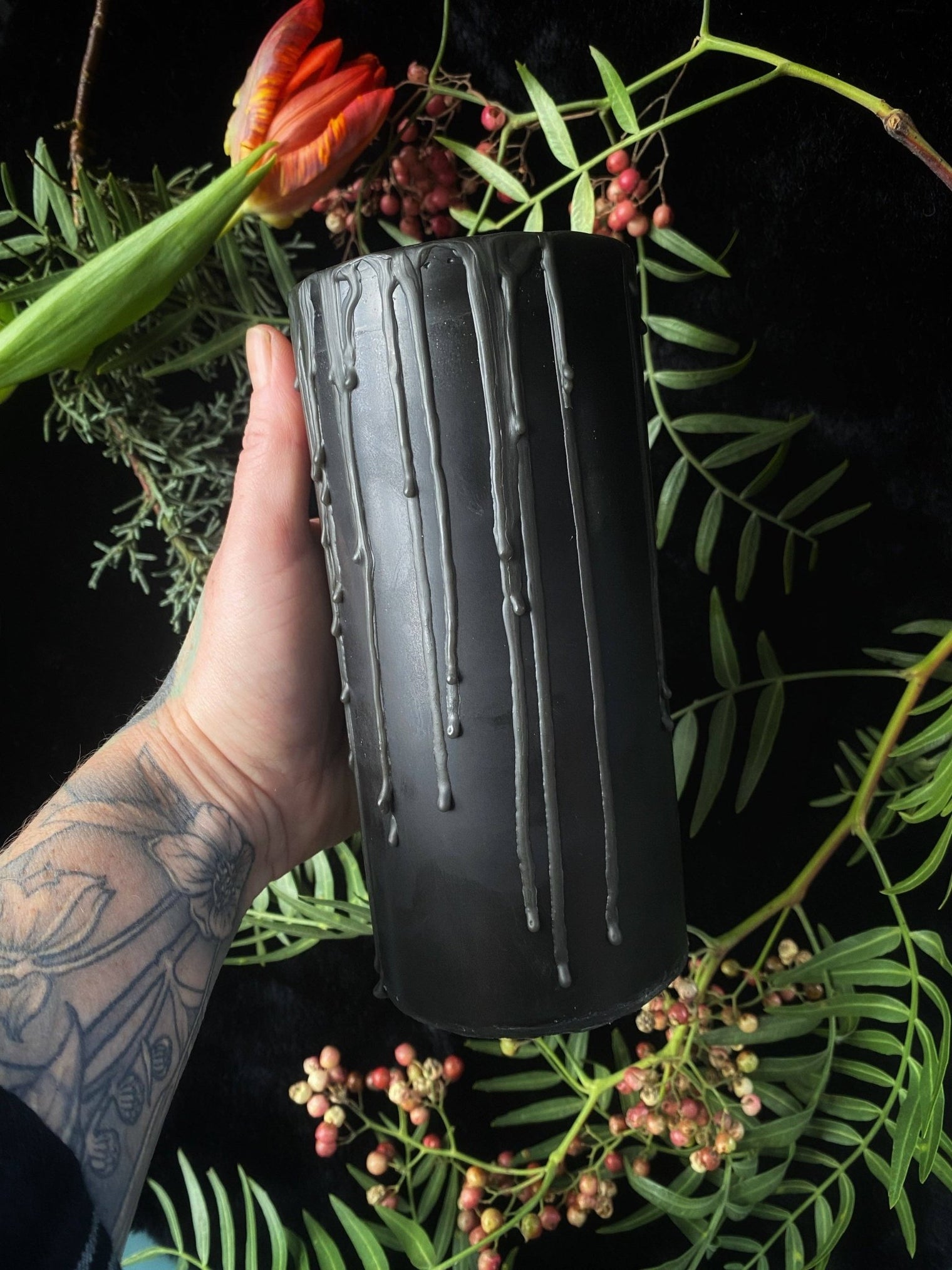 Black textured vase held by a hand with a dark background and greenery