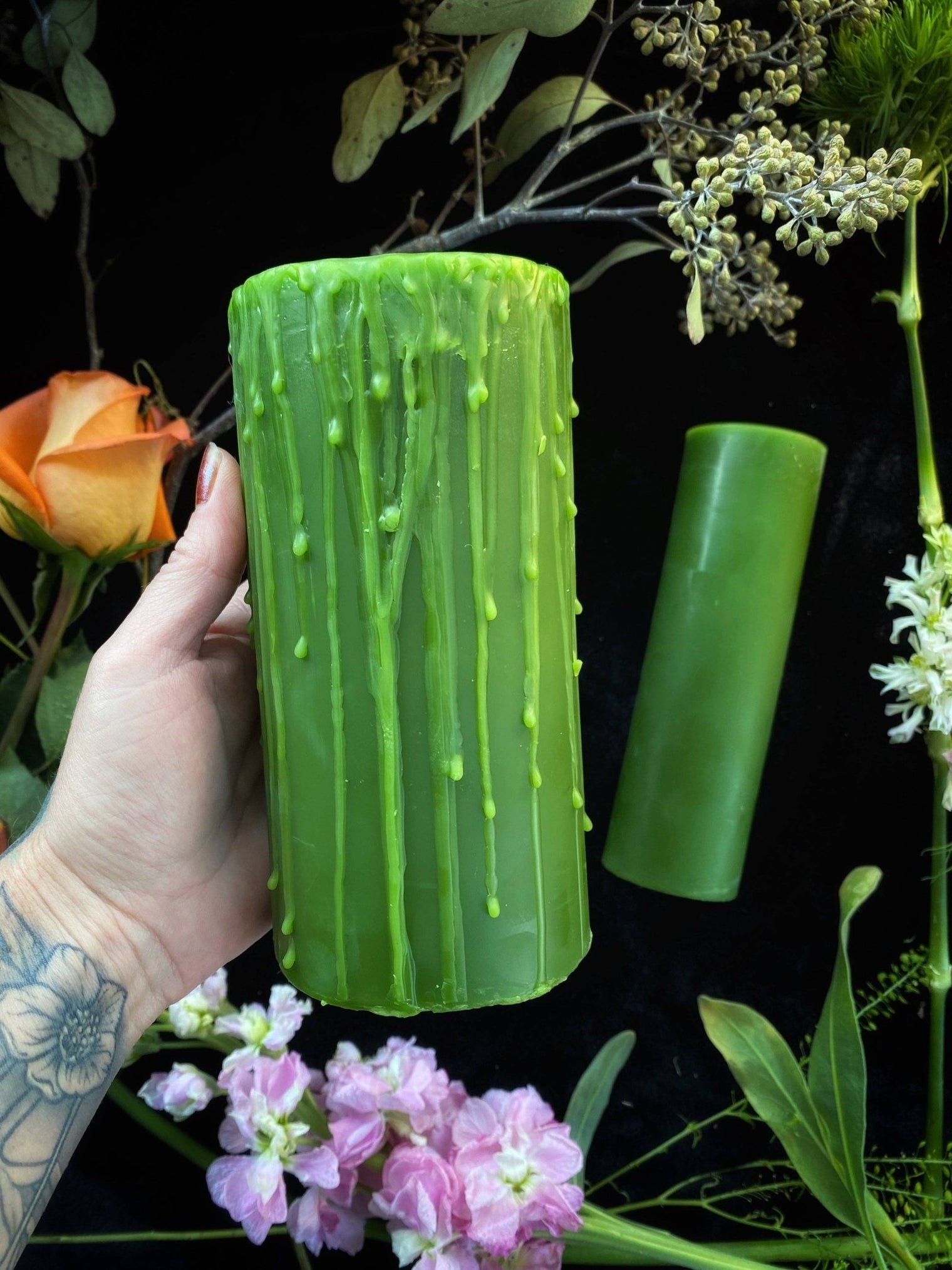 Green textured vase held by a hand with tattoos, surrounded by flowers on a dark background