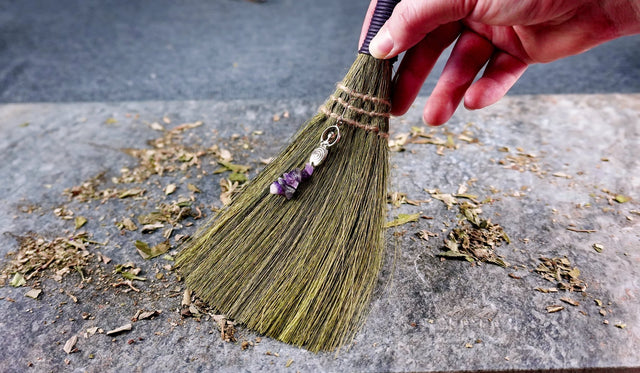 A hand holding an altar broom with a Goddess charm attached, standing on a stone surface surrounded by dried herbs.