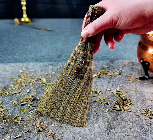 A hand holding an altar broom with a Tree of Life charm, featuring a bundle of natural bristles and a small metal charm.