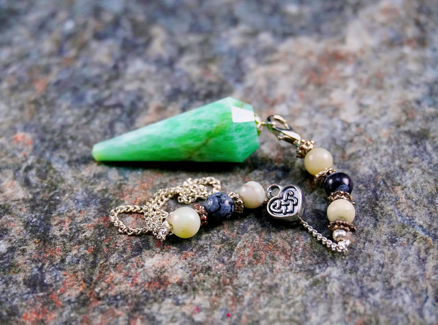 An image of an amazonite hexagonal pendulum with a celtic heart charm and additional beads on a chain, lying on a textured surface.