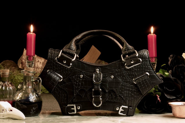 A black bat-shaped handbag with metal buckles, displayed in a dimly lit environment with candles and vintage decorations.