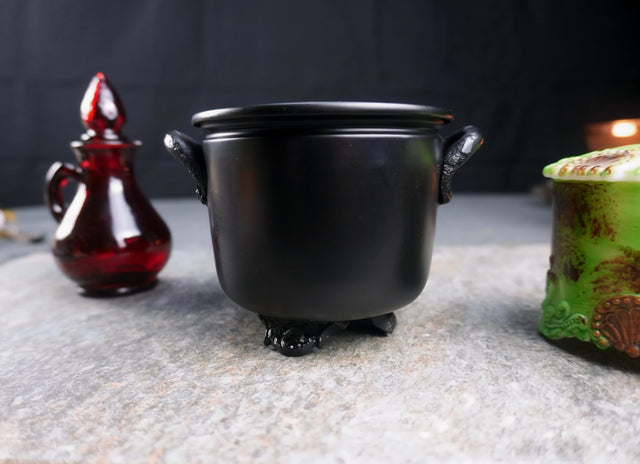 A small matte black metal cauldron with handles, displayed on a stone surface, with a red bottle and a ceramic container in the background.