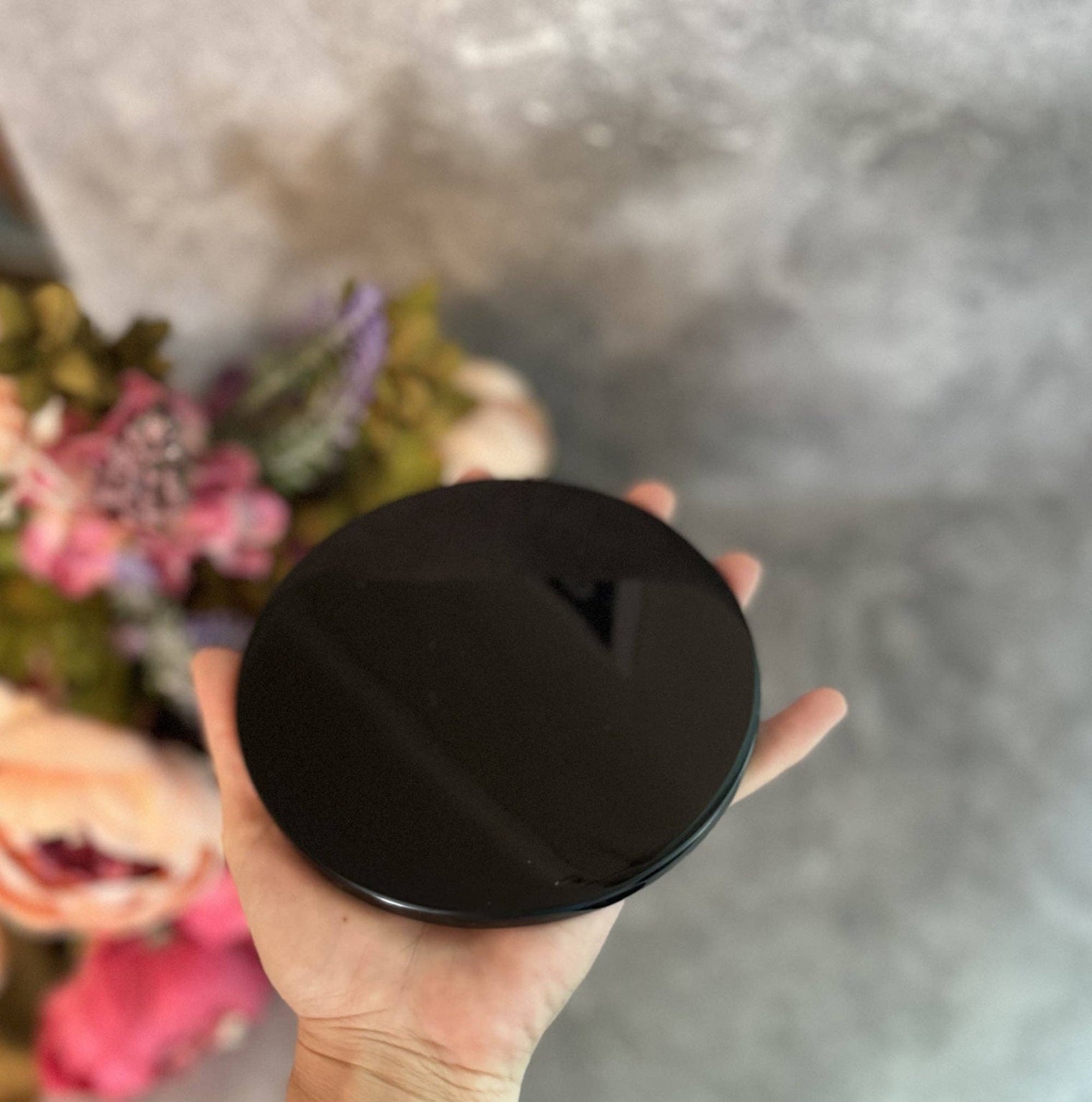 Hand holding a black round object against a blurred background with flowers.