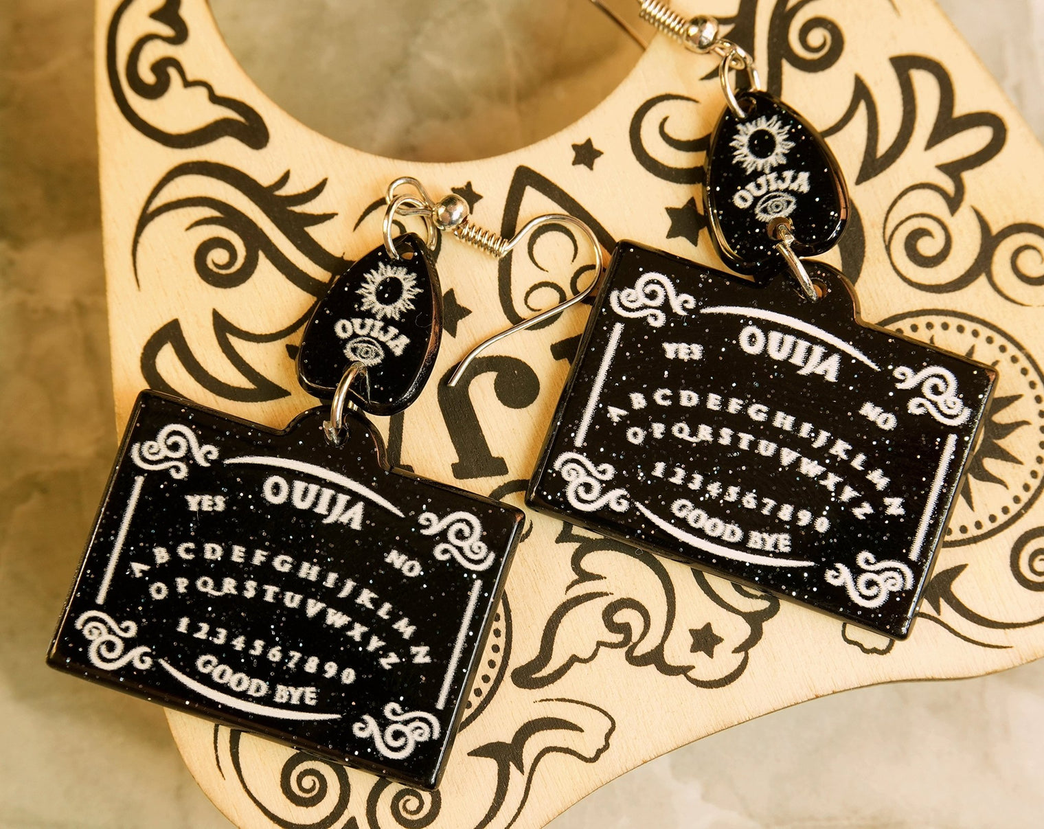 A pair of black Ouija board earrings dangle from a decorative hook, featuring the iconic spirit board design with letters and the planchette.