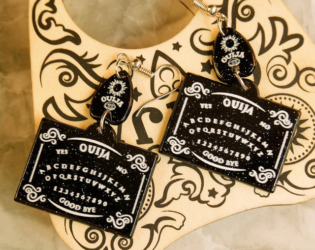 A pair of black Ouija board earrings dangle from a decorative hook, featuring the iconic spirit board design with letters and the planchette.