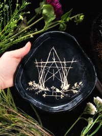 Black embroidered dish with geometric design held by a hand, surrounded by greenery and flowers.