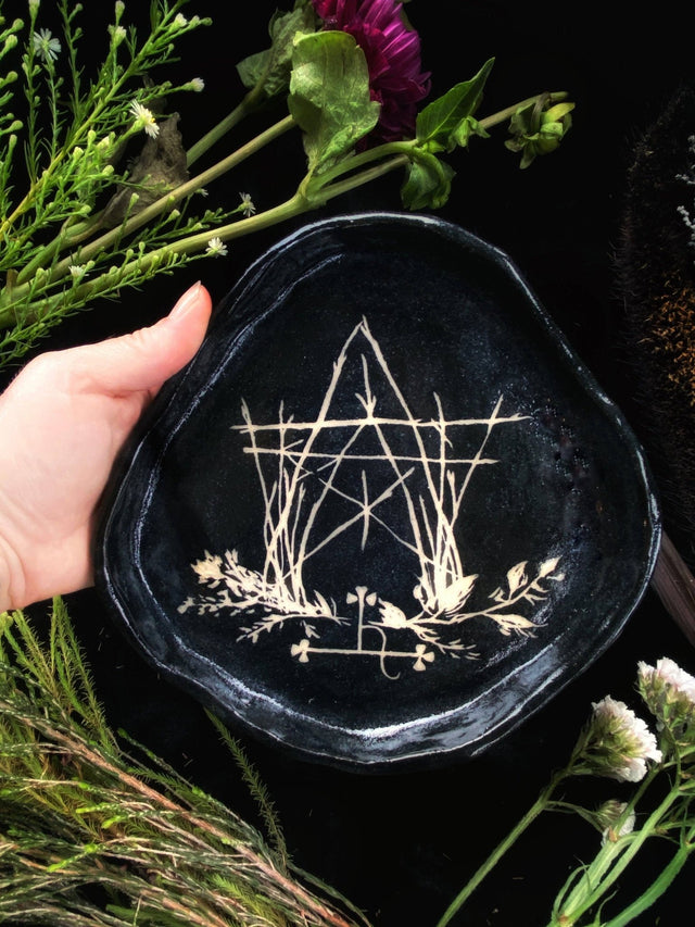 Black embroidered dish with geometric design held by a hand, surrounded by greenery and flowers.