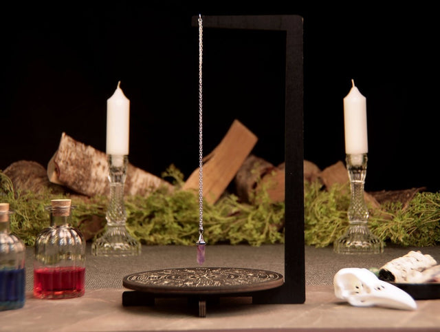 A divination pendulum stand with various symbols including zodiac signs and letters, with a pendulum hanging from the top and candles in the background.