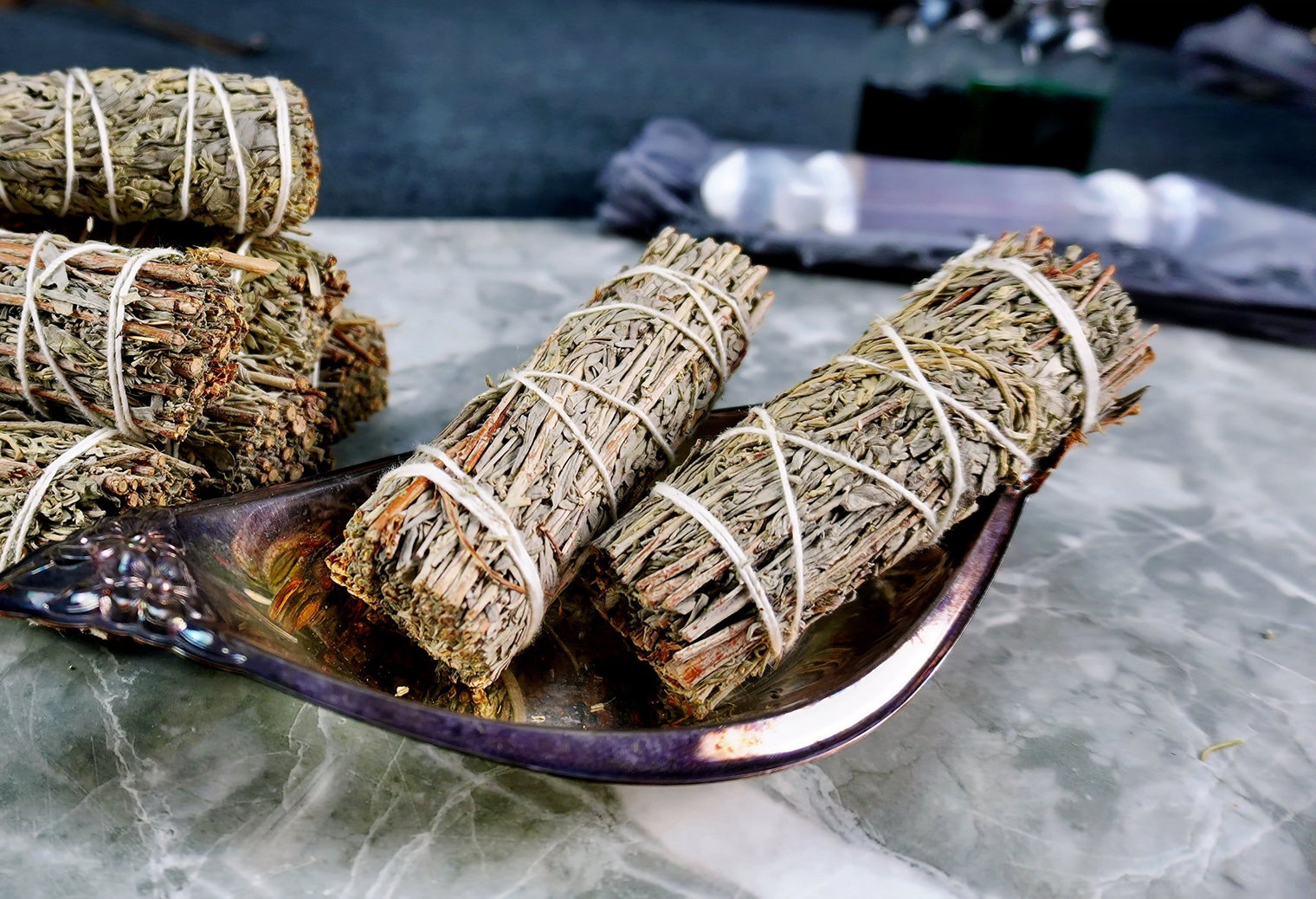 Bundles of Blue Sage Smudge Sticks tied with string, placed on a metal dish on a marble surface.