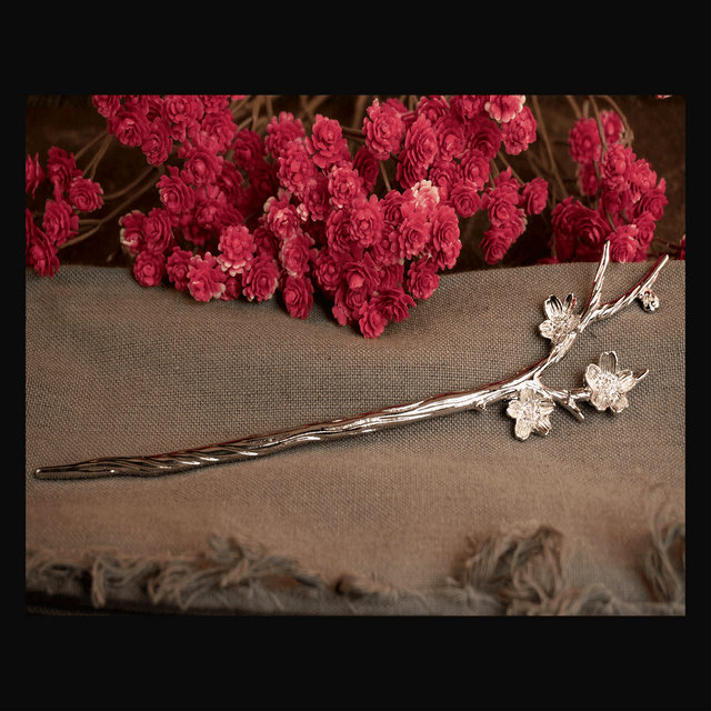 A silver-colored metal hair pin designed in the shape of a branching twig with flowers, displayed against a fabric background with red floral arrangement.