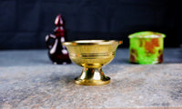 A polished brass ritual drinking bowl displayed on a stone surface with a blurred bottle and container in the background.