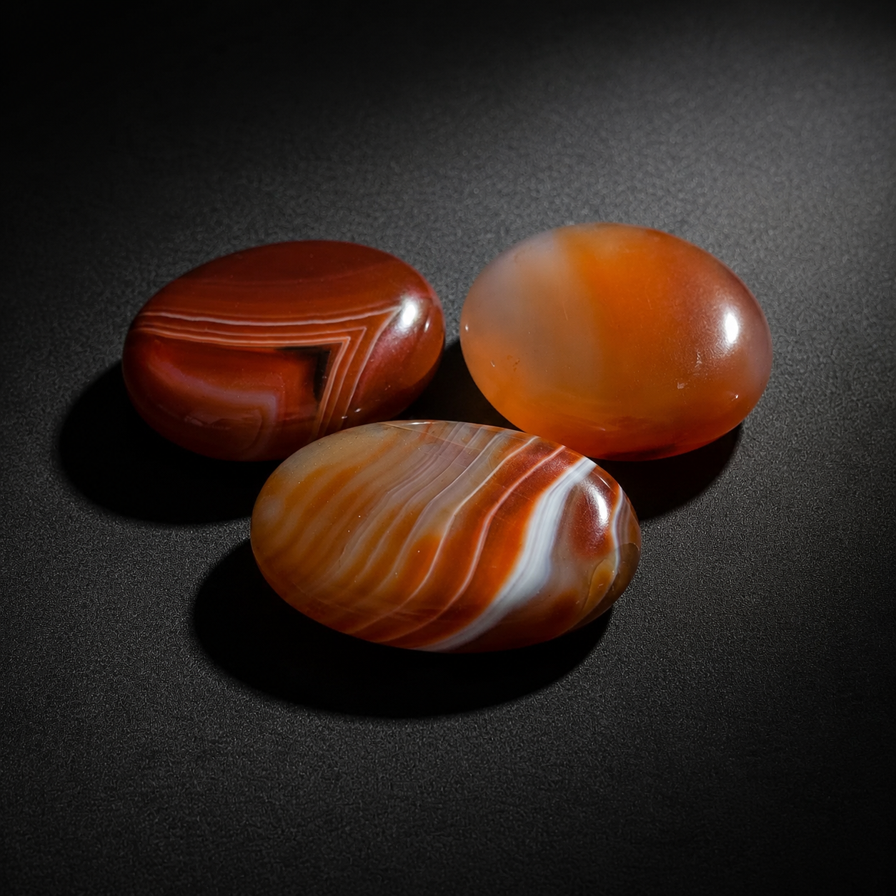 Three polished orange-red carnelian palm stones with natural banding on a matte black surface