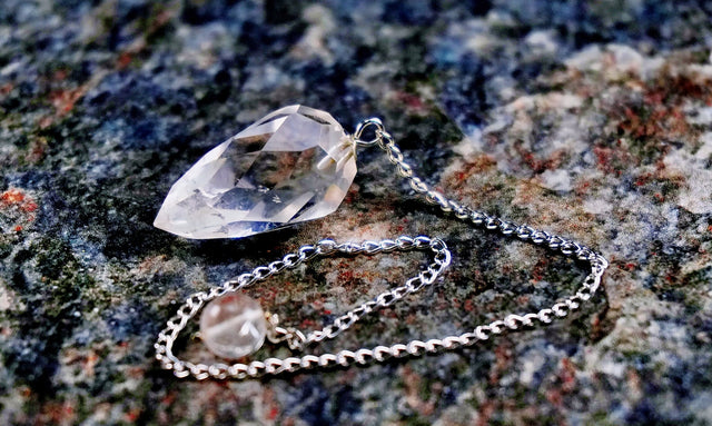 A clear quartz crystal pendulum with a silver chain, resting on a textured surface.