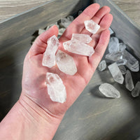Hand holding clear crystal stones with a tray of additional stones below on a wooden surface.
