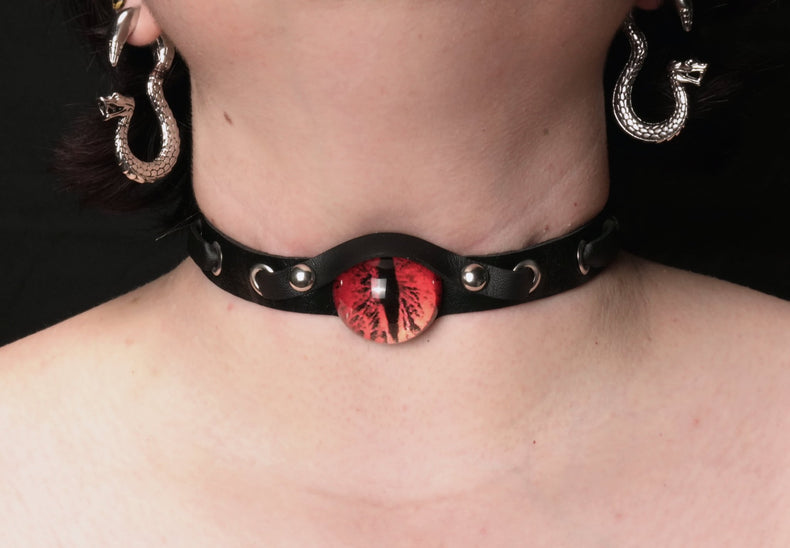 A close-up image of a person wearing a black leather choker collar with a red demon eye at its center.