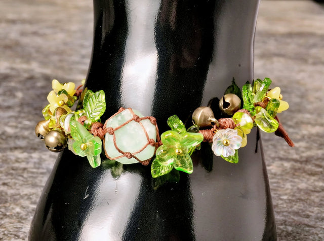 A beaded bracelet with green flower and leaf designs, accented by butterflies and bells, with a gemstone in the center encased in a woven cage.