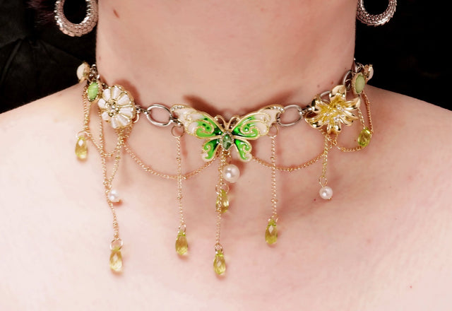 A close-up image of a green butterfly necklace with tassels, featuring layered gems and a gold-colored chain.