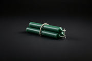 Green chime candles tied with gold string, lit at one end, on a dark background with a copper bowl and herbs.