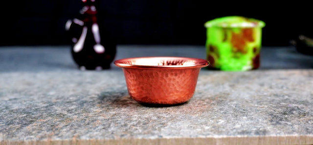 A hammered copper ritual bowl with a warm, textured finish, displayed on a stone surface, with a bottle and a green container in the background.