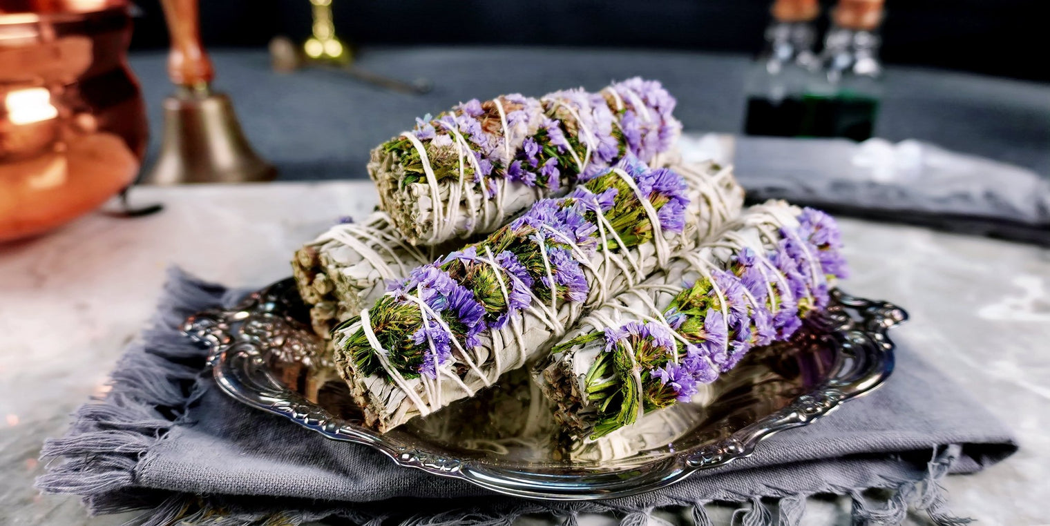 Three smudge sticks made from sage and adorned with lilac statice and sinuata blooms, displayed on a metal tray on a gray background.